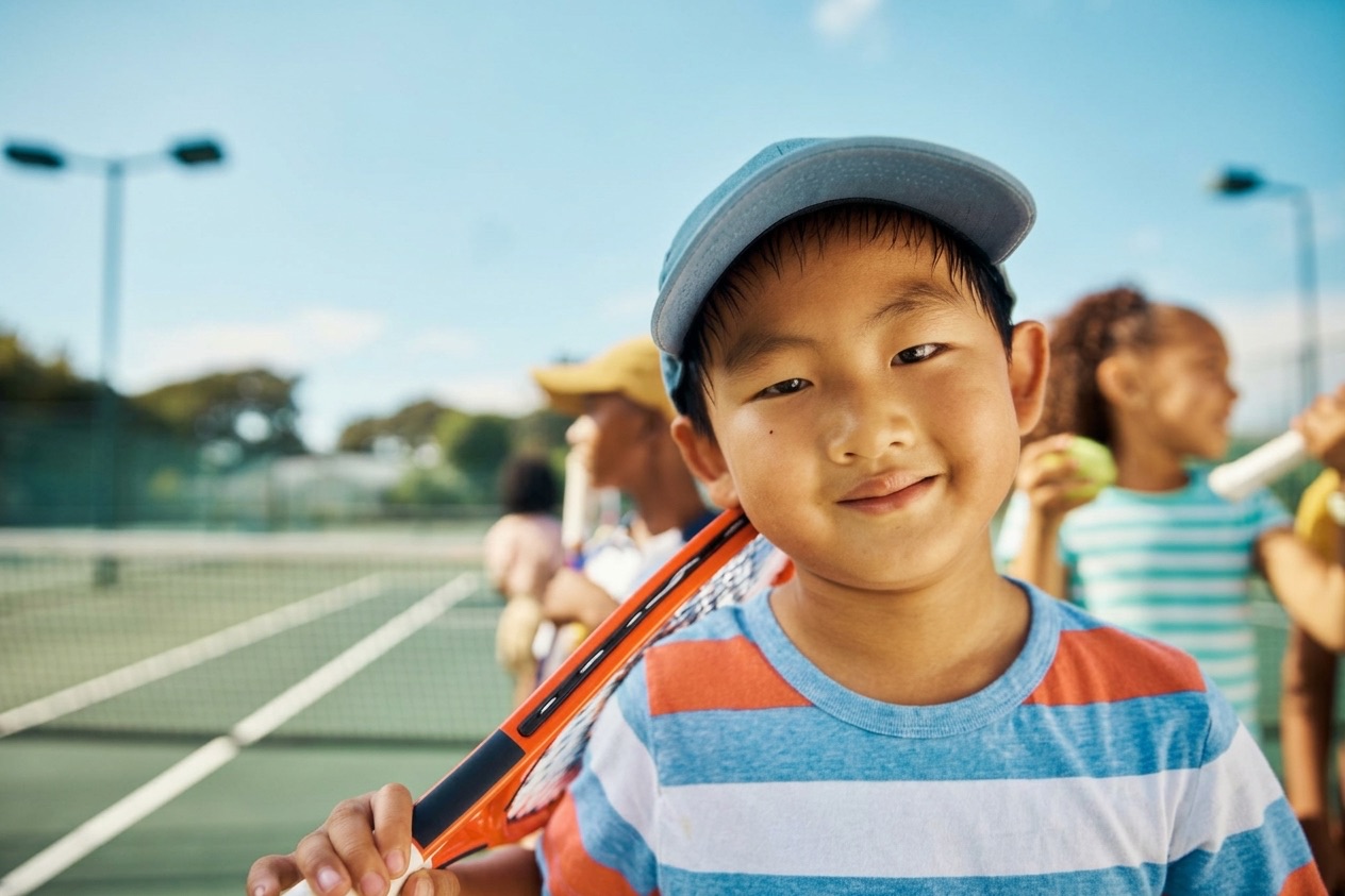 A smiling boy in a striped shirt and cap rests a racket on his shoulder on a sunny court